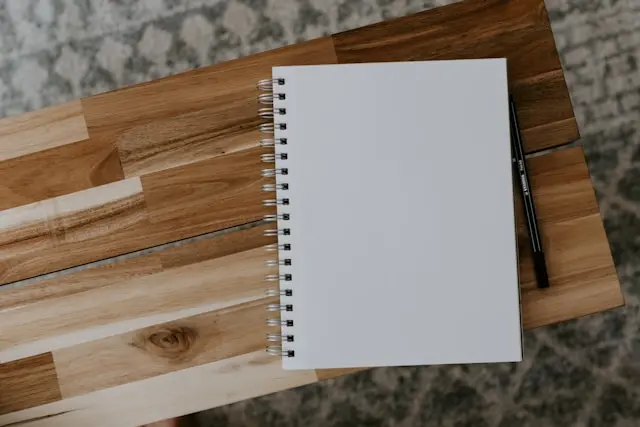 Caderno de espiral em branco sobre tábua de madeira, com caneta preta ao lado, pronto para anotar como fazer um resumo de um livro.